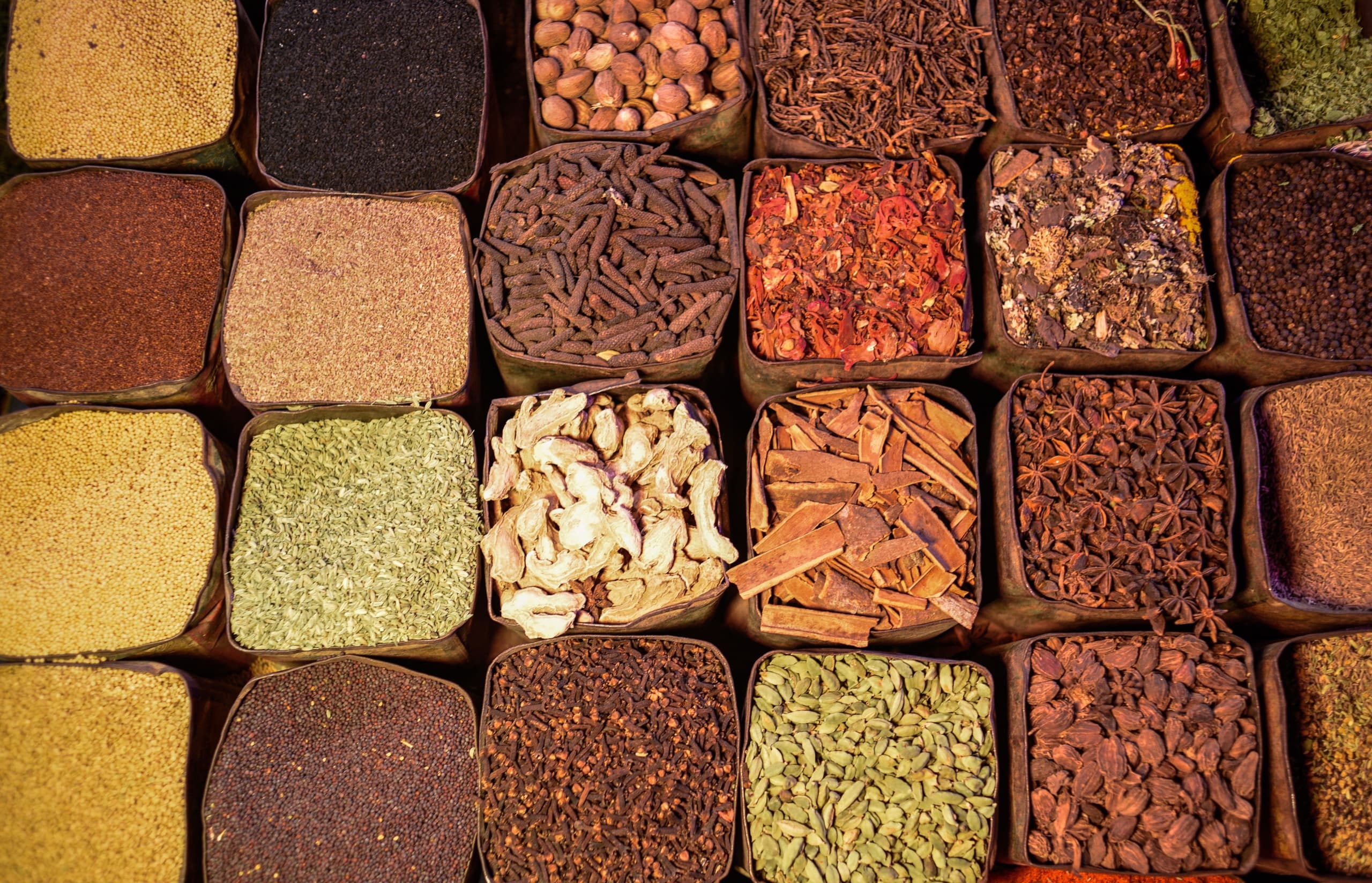 Dried spices bulked side by side in square bins: cinnamon quills, star anise, cardamom, cloves, dried ginger and peppercorns