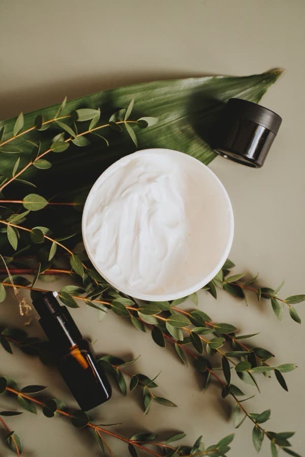 An open jar of cream with eucalyptus sprigs and a glass dropper